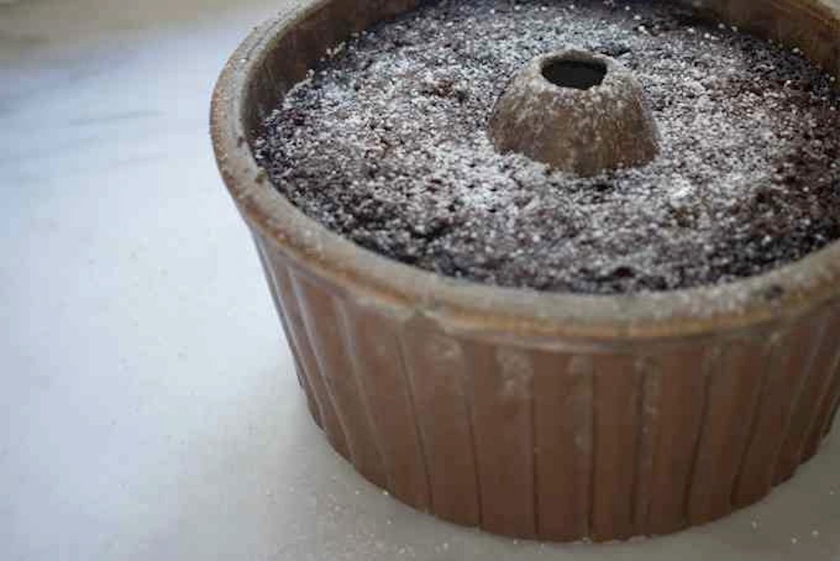 A chocolate cake dusted with powdered sugar sits in a brown fluted bundt pan on a white surface.