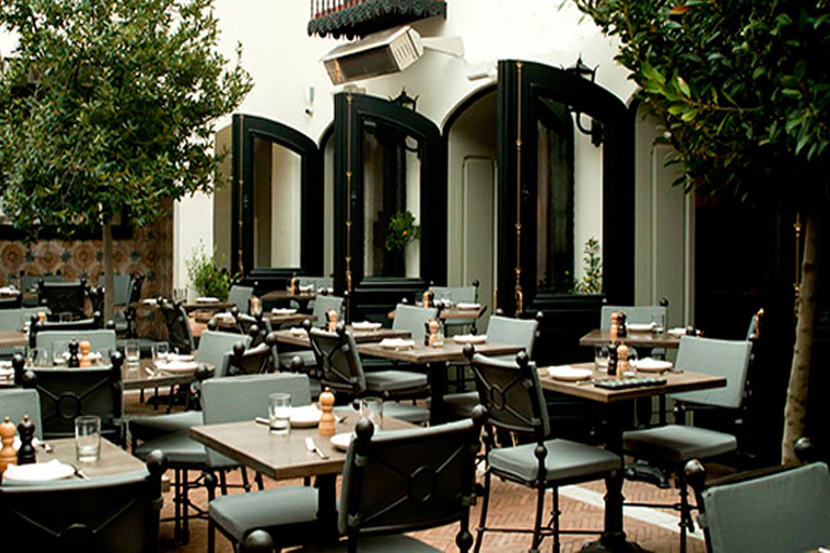 Outdoor restaurant patio with empty tables and chairs arranged neatly. The area is surrounded by greenery and large windows with open black frames, creating a welcoming and elegant atmosphere.