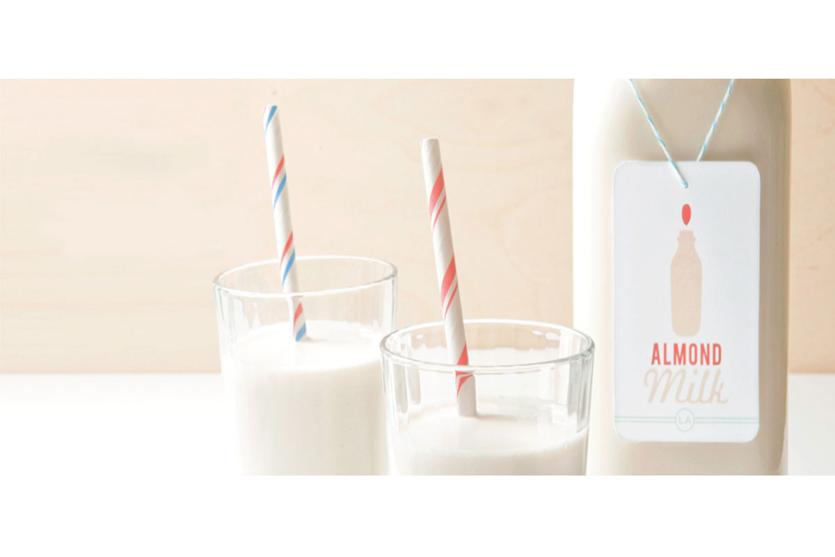 Two glasses of almond milk with striped straws sit next to a labeled bottle of almond milk on a light-colored surface.
