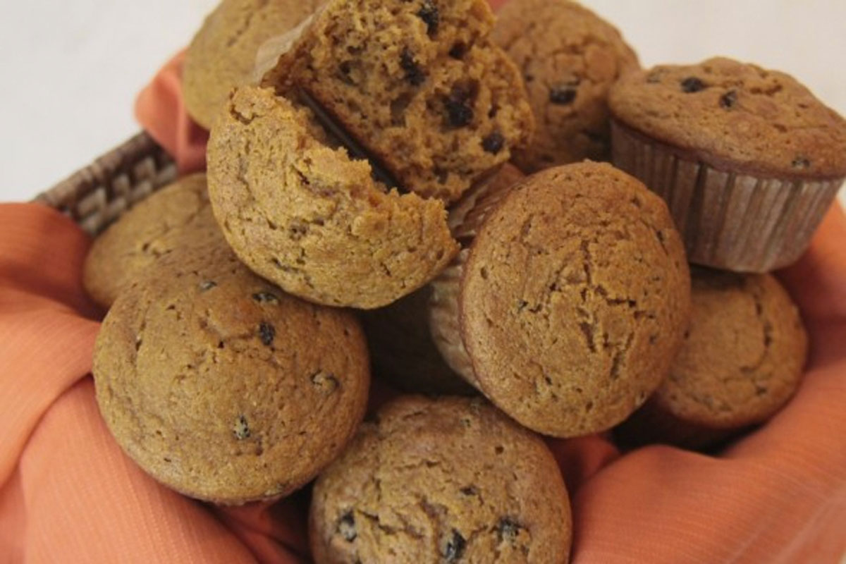 A basket lined with an orange cloth holds several pumpkin muffins with chocolate chips, with one muffin broken open to show the inside texture.