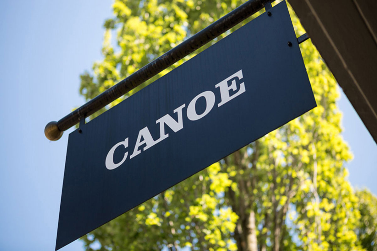 A dark blue rectangular sign with the word CANOE in white capital letters hangs outside, with green leafy trees and blue sky in the background.