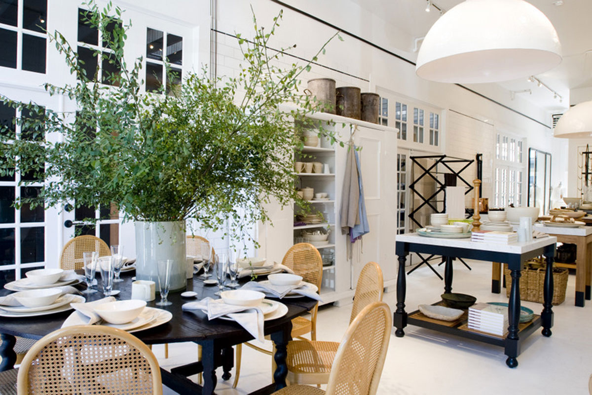 A bright, modern dining room with a round dark wood table set for six, wicker chairs, a large vase with green branches, open shelves with dishes, and a black rolling cart in a white, airy space.