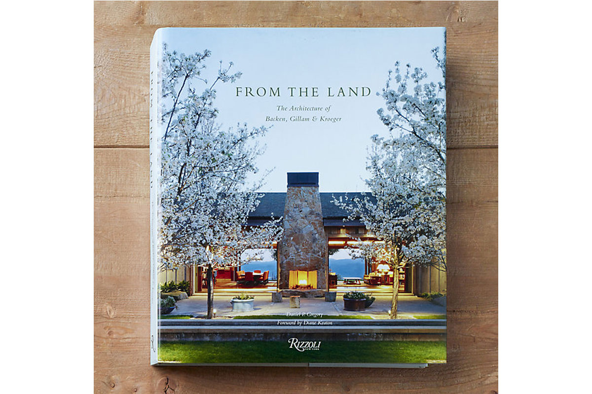 A hardcover book titled From the Land featuring a house with large windows, a stone chimney, and blooming trees on the cover, placed on a wooden surface.