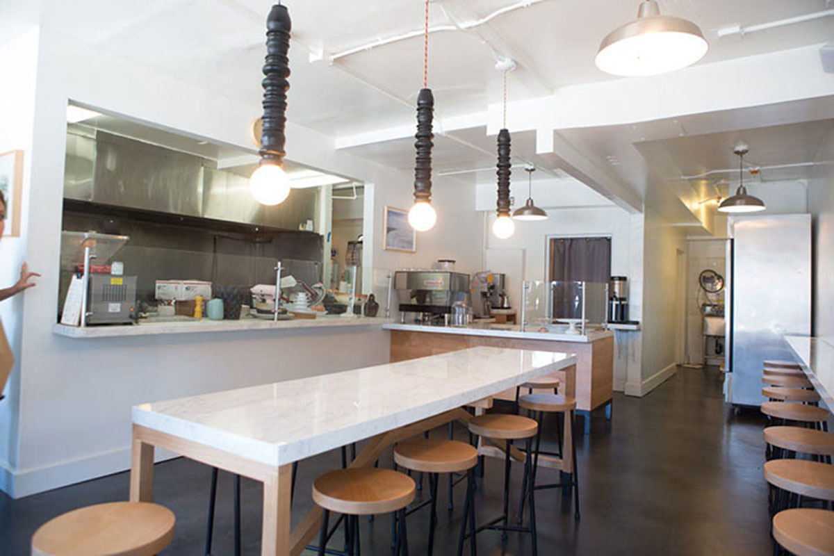 A modern, minimalistic cafe interior with a long marble-topped communal table, wooden stools, hanging pendant lights, an open kitchen, and coffee machines on the counter. The space is bright and clean.
