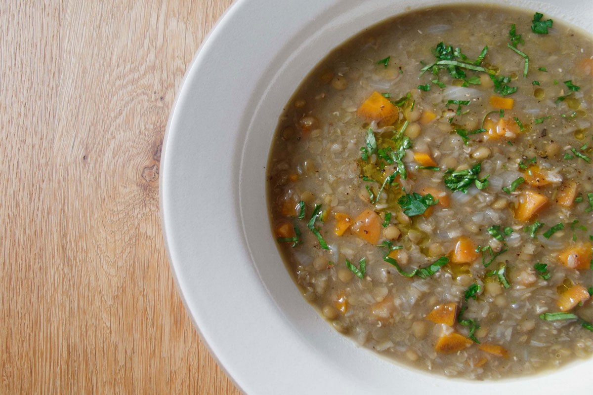 A white bowl filled with lentil soup containing diced carrots and topped with chopped parsley, placed on a wooden surface.