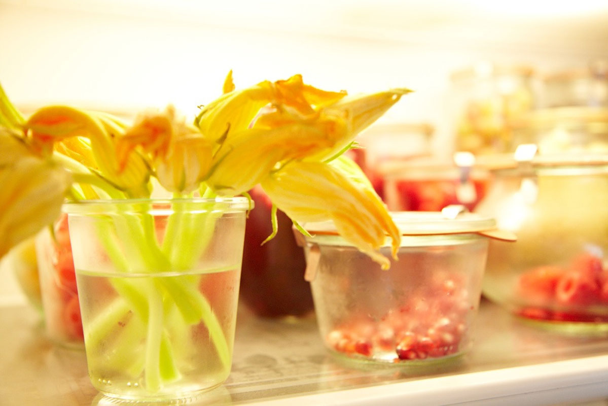 A close-up of squash blossoms in a glass of water inside a refrigerator, surrounded by jars of preserved food and various other items on the shelf.