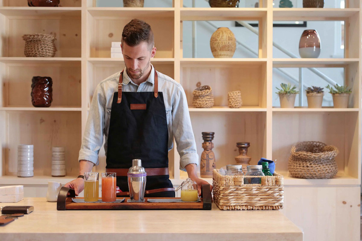 A bartender wearing a dark apron stands behind a light wooden counter, preparing drinks on a tray. Woven baskets, pottery, and succulents are displayed on shelves in the background.