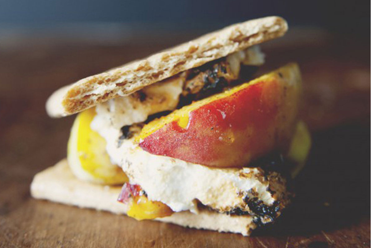 A close-up of a smore made with graham crackers, roasted marshmallow, and slices of grilled peach, set on a wooden surface.