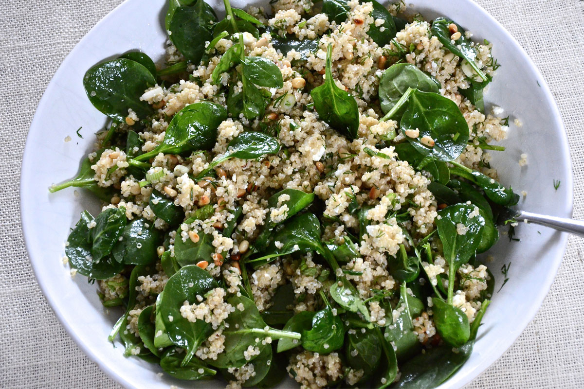 A bowl of quinoa salad with fresh spinach leaves, chopped herbs, and nuts, served on a light-colored cloth with a spoon partly visible on the side.