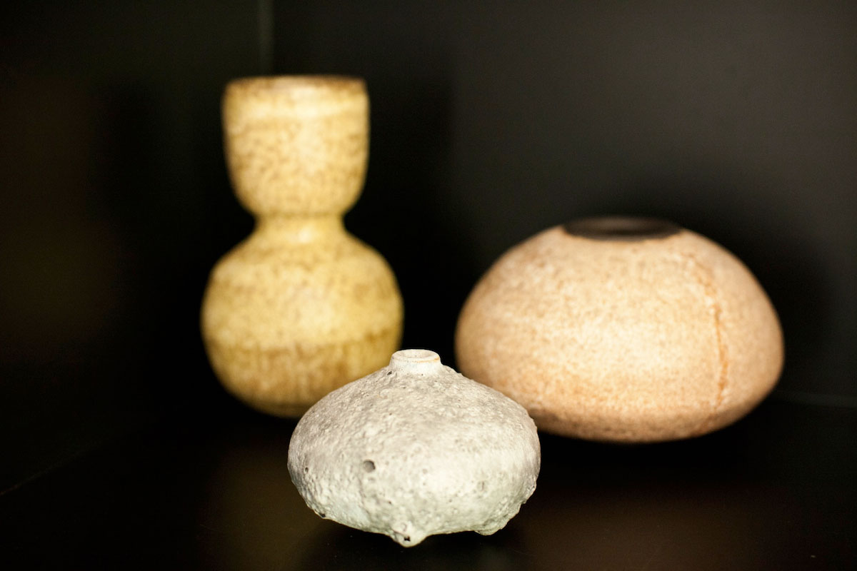 Three textured ceramic vessels on a black background: a tall, yellow double-gourd-shaped vase; a round, tan vessel; and a small, light gray, flattened bottle with a narrow opening in front.