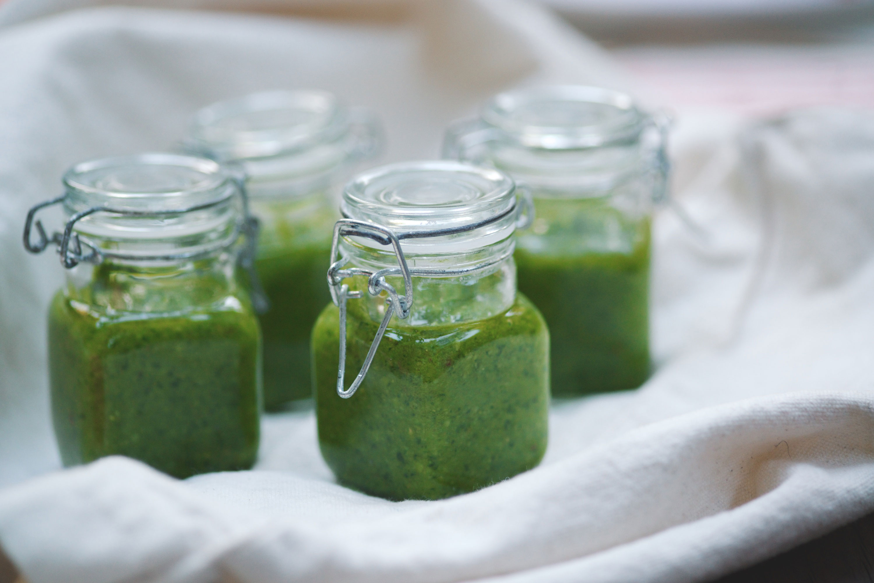 Four small glass jars with metal clasps filled with green sauce or pesto are placed on a white cloth, creating a fresh and inviting presentation.