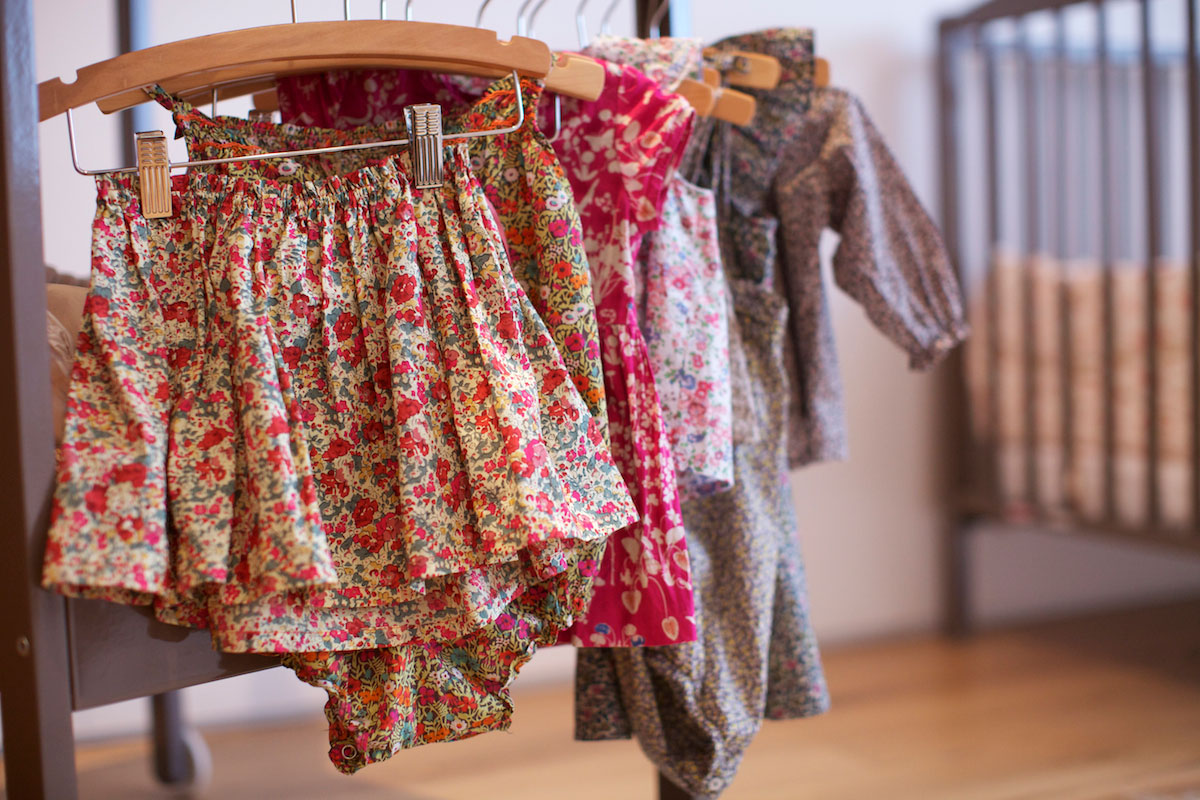 Several colorful, floral-patterned toddler dresses and skirts are hanging on wooden hangers, with a crib visible in the blurred background. The clothes are neatly arranged in a warm, cozy nursery room.