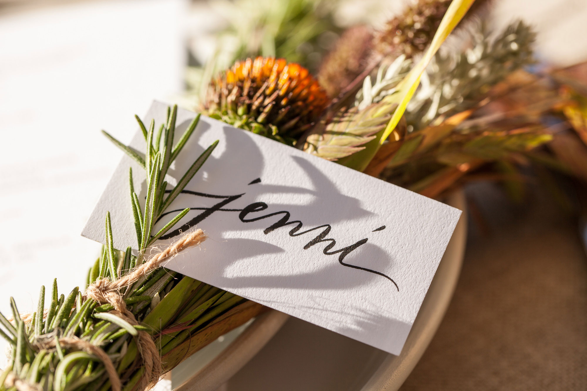 A place card with the name Jenni in cursive rests on a bundle of herbs and dried flowers, tied with twine, atop a plate in warm sunlight.