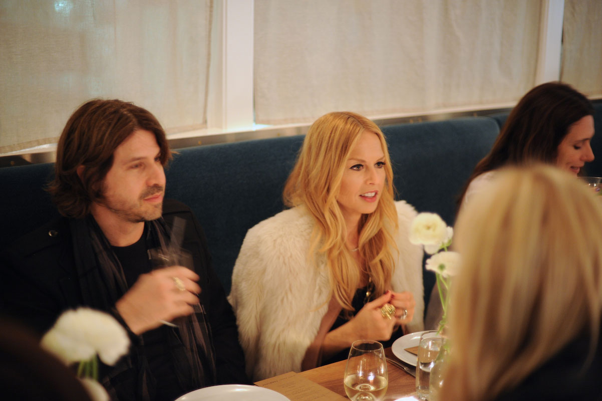 Two people sit at a table in a restaurant, engaged in conversation. The woman wears a white fur coat, and the man has long brown hair and holds a drink. White flowers and plates are arranged on the table.