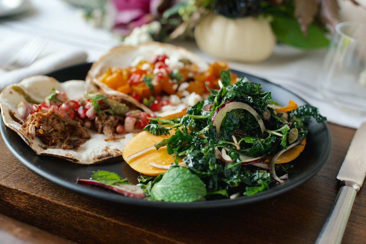 A black plate with two tacos filled with shredded meat, salsa, and vegetables, alongside a fresh salad of kale, thinly sliced yellow beets, red onions, and herbs.