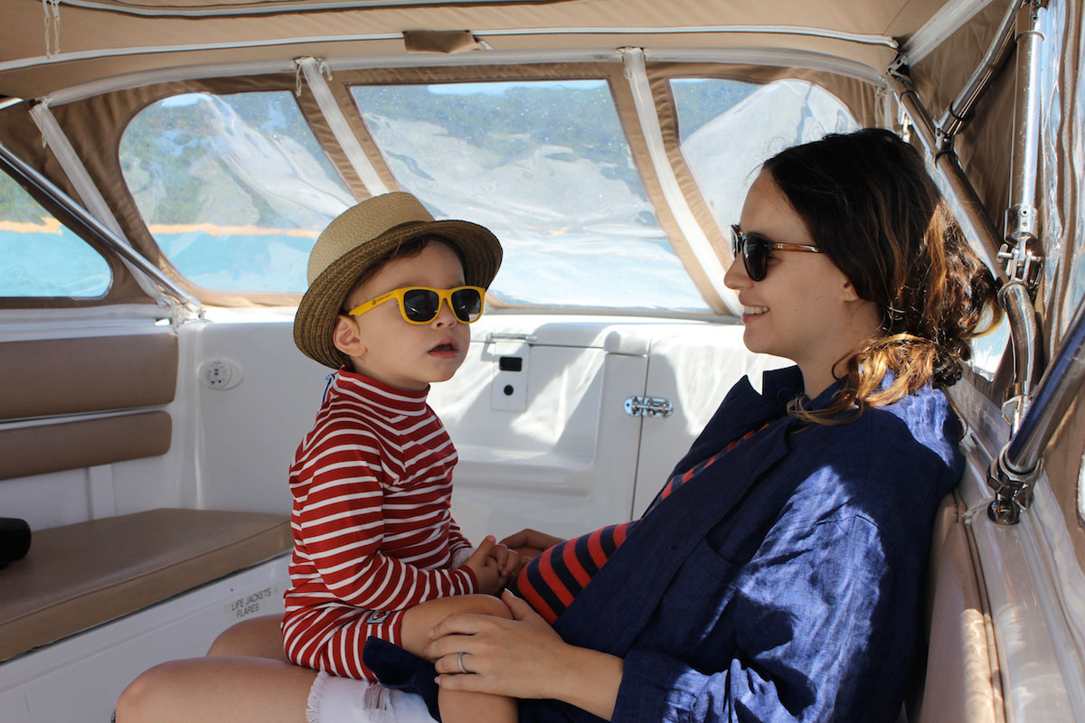 A woman and a young child wearing sunglasses sit together on a boat. The child wears a hat and a red striped shirt, while the woman smiles and wears a blue jacket. Bright sunlight and water are visible outside.