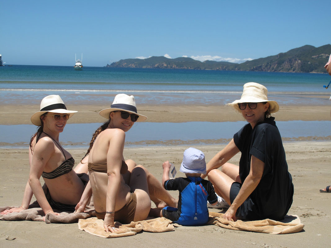 Three women in hats sit on a sandy beach with a young child in a blue outfit, all facing the camera. The ocean and distant hills are visible in the background under a clear sky.