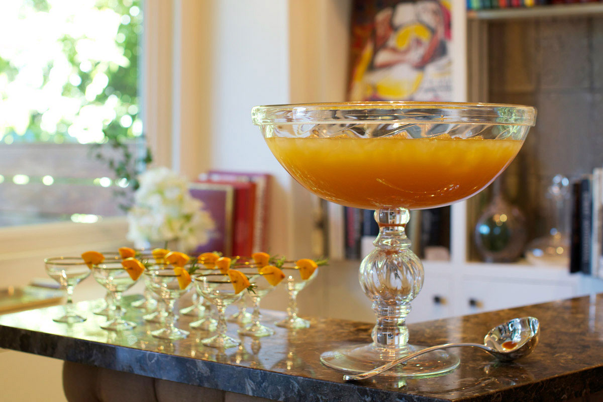 A large glass punch bowl filled with orange punch sits on a marble counter beside several small glasses garnished with orange slices; a metal ladle rests nearby. The background includes books and a window.