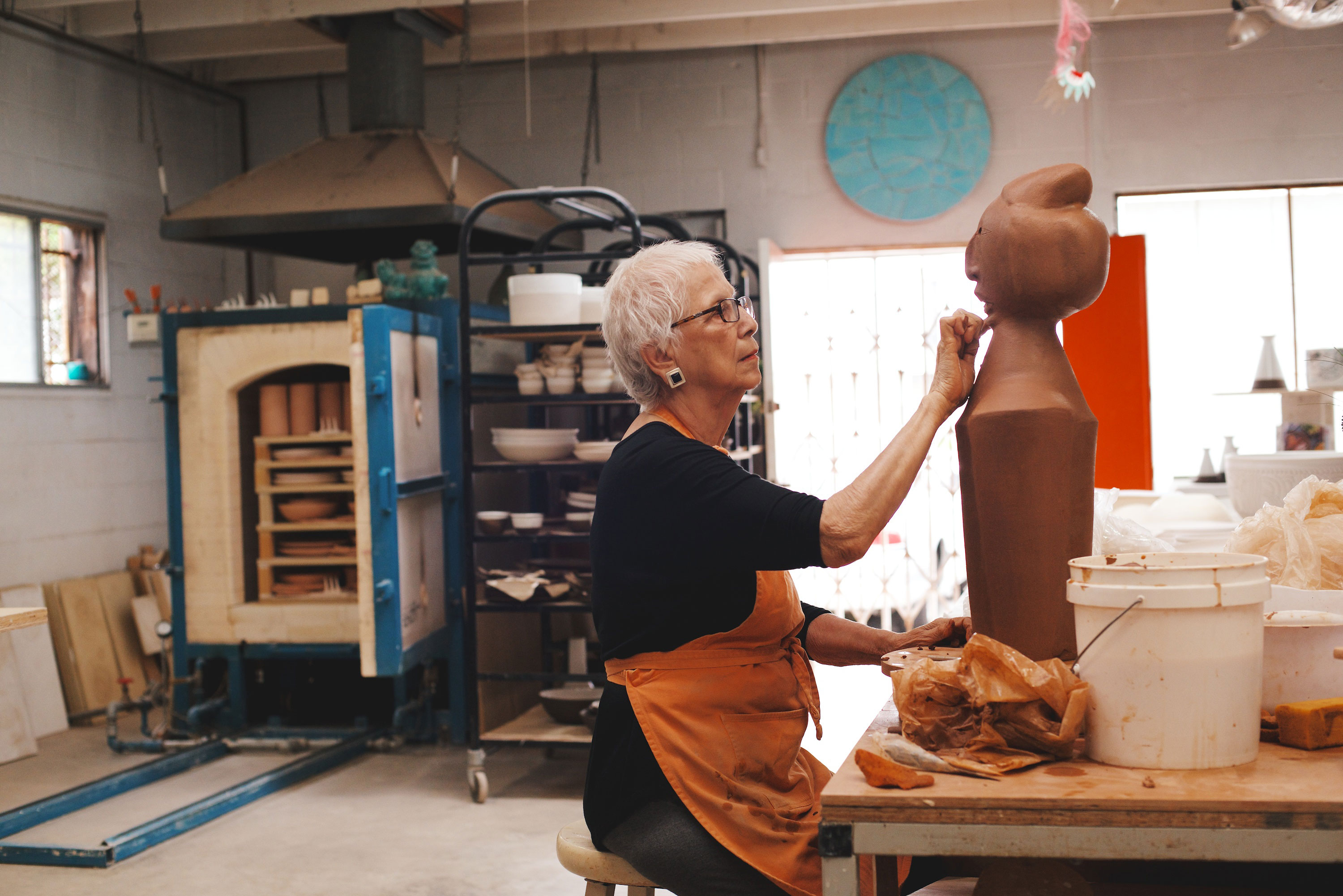 An older woman wearing glasses and an apron shapes a large clay sculpture in a pottery studio, surrounded by ceramics, a kiln, tools, and shelves filled with pottery items.