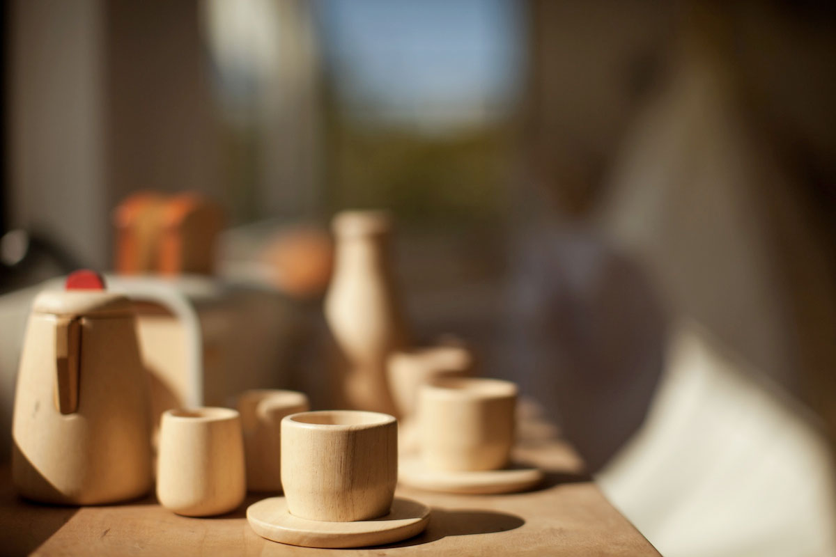 A close-up of wooden toy kitchenware, including cups, saucers, a teapot, and a bottle, arranged on a wooden surface with soft natural light and a blurred background.