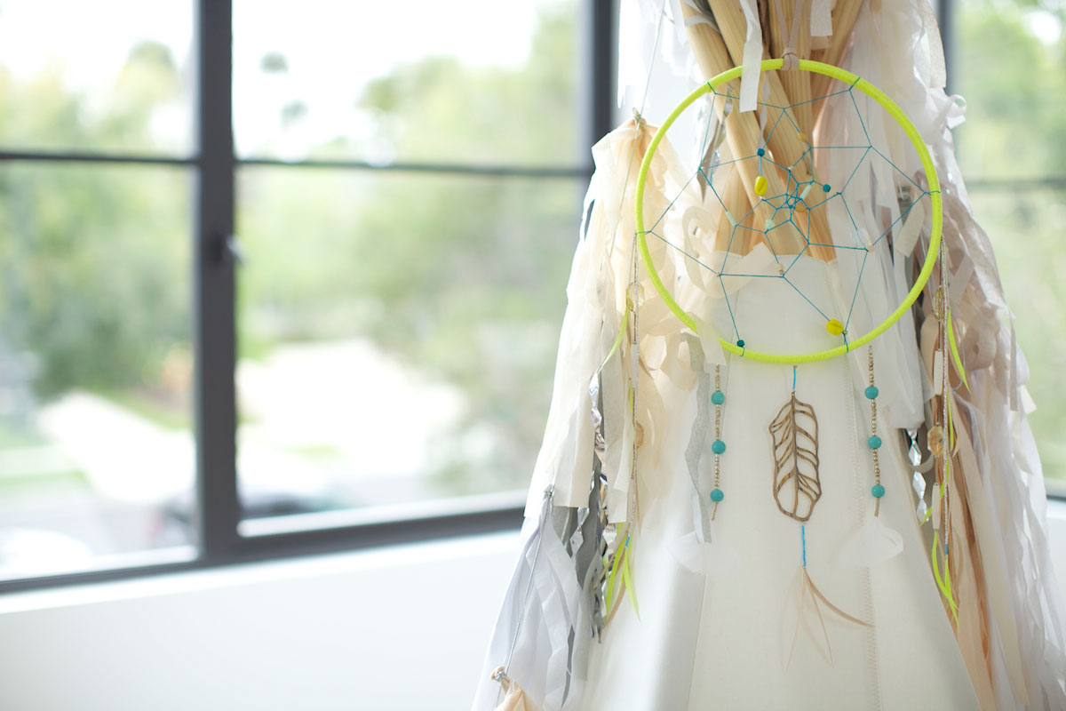 A neon green dreamcatcher with blue beads and feather charms hangs on a white fabric structure near a window, with soft natural light and greenery visible outside.