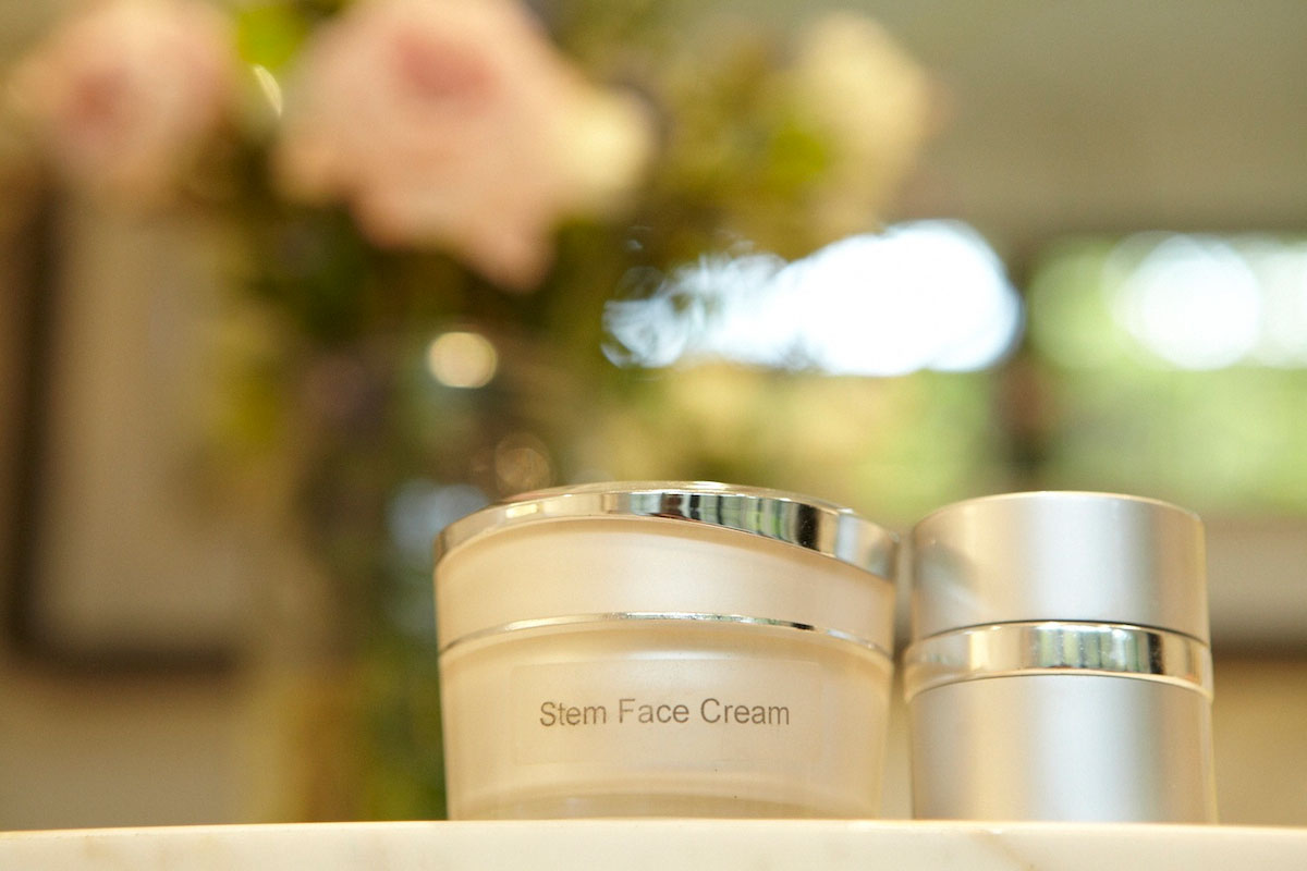 Two jars of skincare products, one labeled Stem Face Cream, sit on a surface with a blurred background of flowers and greenery.