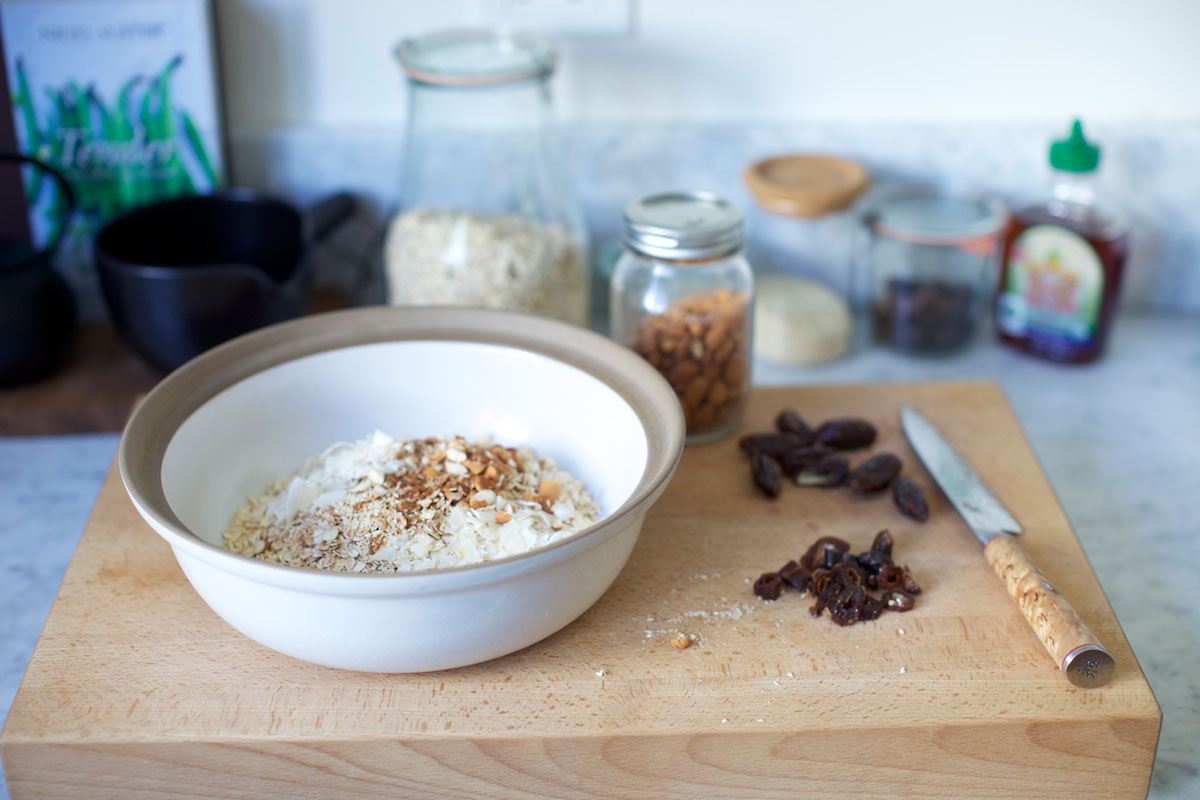 A bowl with oats and chopped nuts sits on a wooden cutting board next to sliced dates and a knife. In the background are jars of oats, almonds, other ingredients, and kitchen items on a counter.