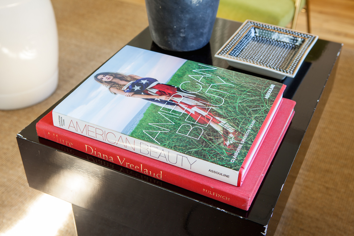 Two coffee table books are stacked on a black table. The top book, American Beauty, features a woman with an American flag. The bottom book is Diana Vreeland. Nearby are a potted plant and a square decorative tray.
