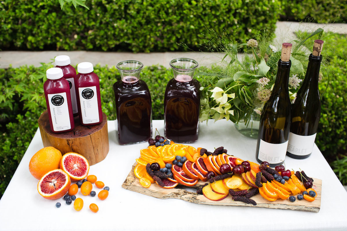 A table set outdoors with fresh fruit juice bottles, two pitchers of dark juice, two wine bottles, a rustic board with sliced oranges, berries, cherries, and assorted greenery in the background.