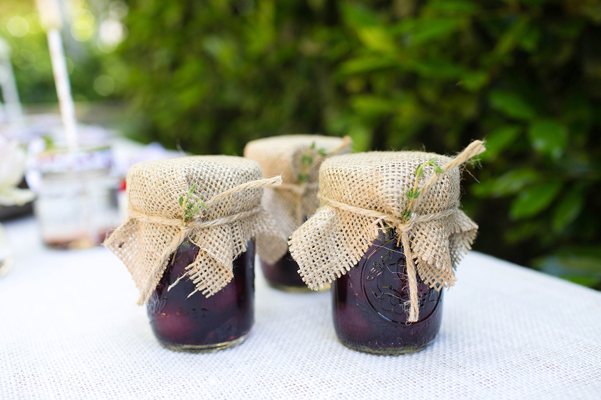 Three glass jars filled with dark jam are covered with burlap fabric and tied with string, sitting on a white tablecloth outdoors, with green foliage in the background.
