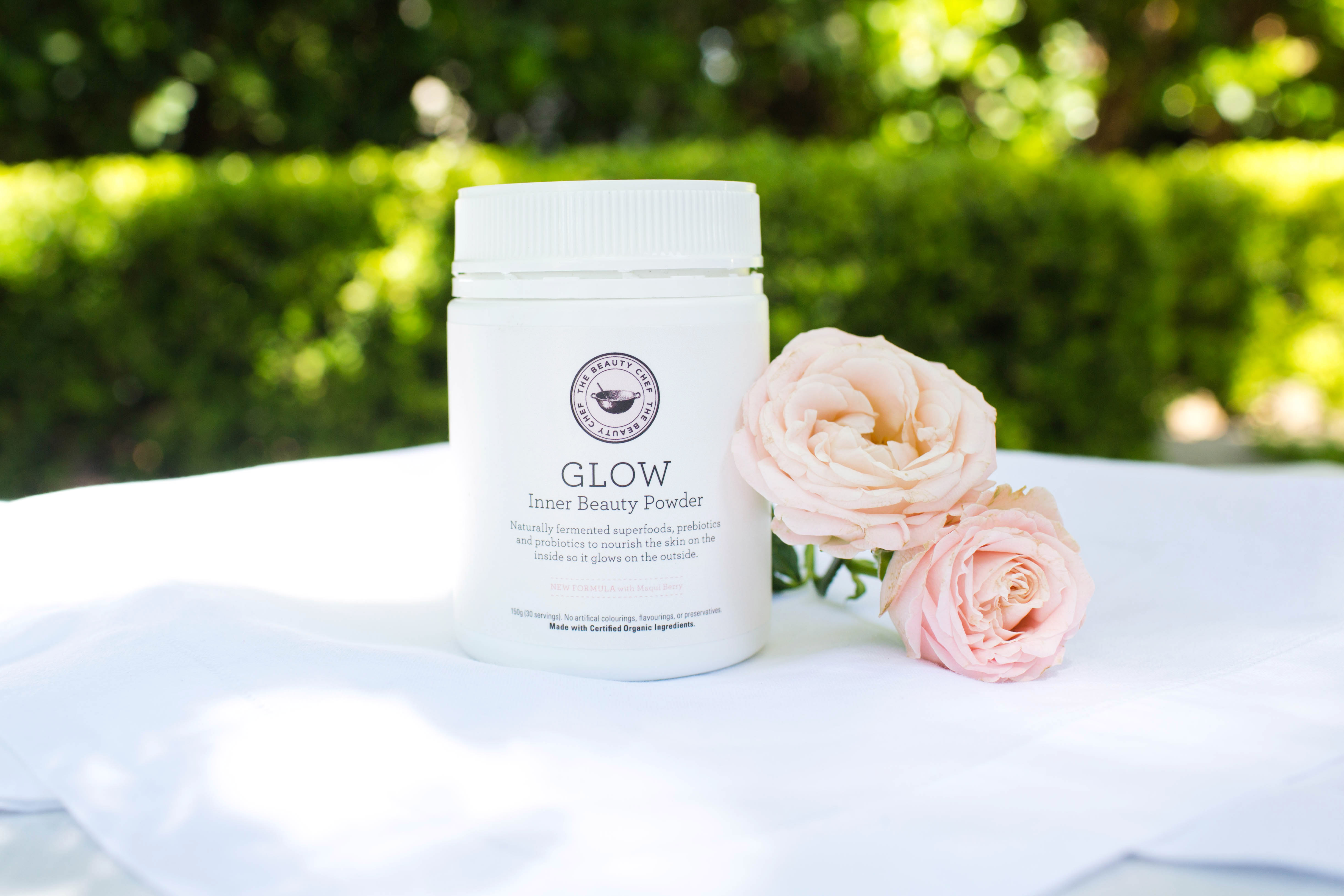 A container of GLOW Inner Beauty Powder sits on a white cloth next to two pale pink roses, with a green leafy background and soft sunlight illuminating the scene.