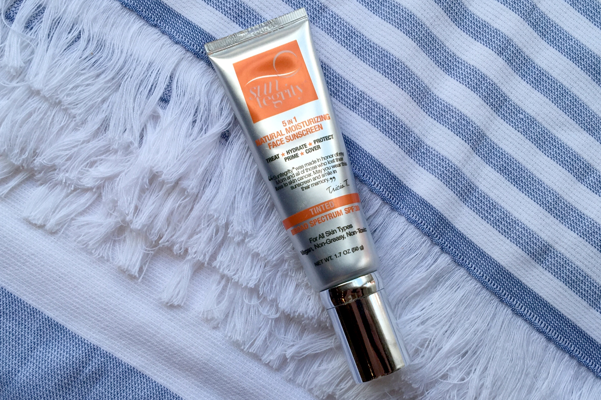 A tube of Suntegrity 5-in-1 Natural Moisturizing Face Sunscreen rests on a white and blue striped towel with fringe detailing.
