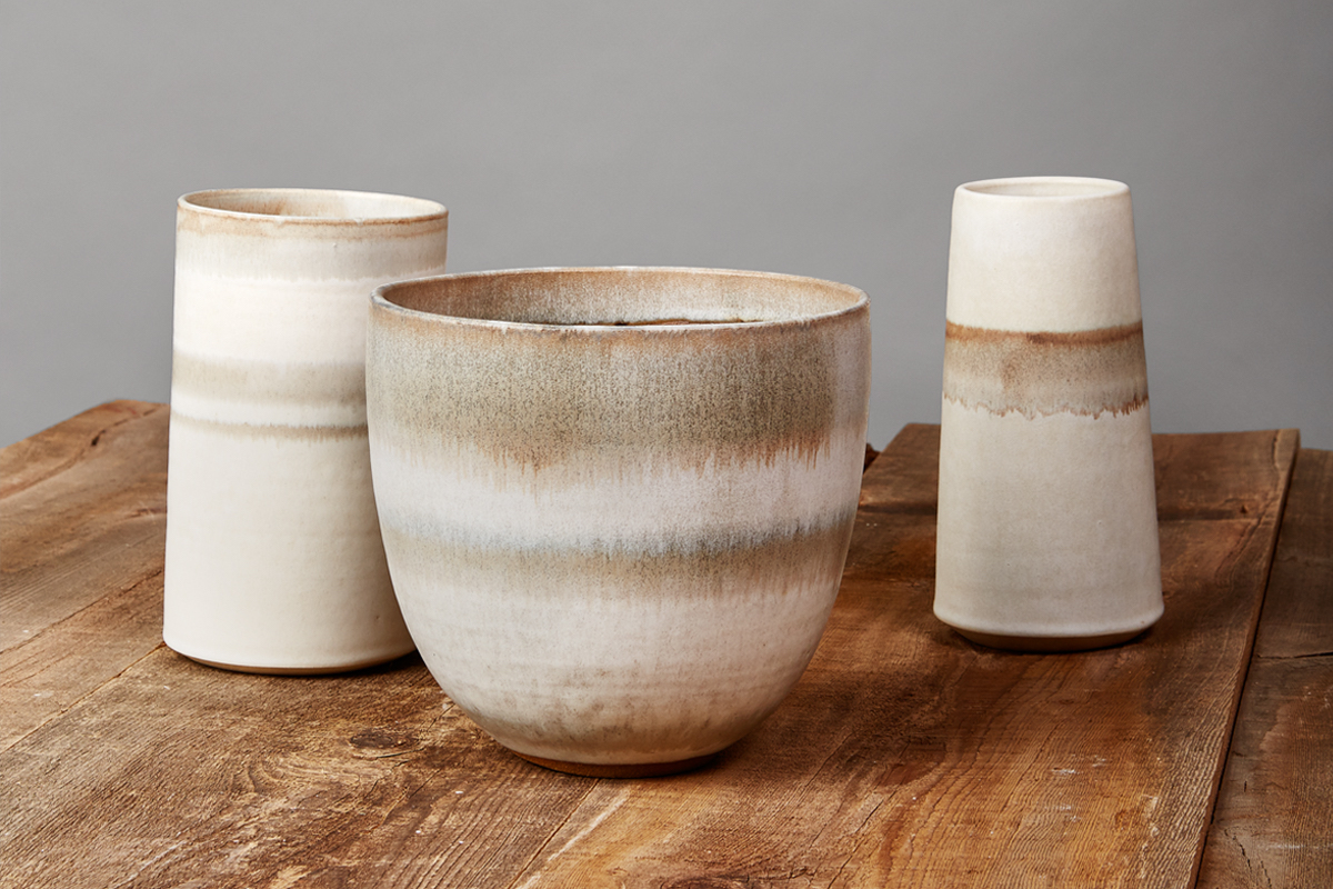 Three ceramic vases with earthy, gradient tones of beige, brown, and cream sit on a rustic wooden table against a plain gray background.