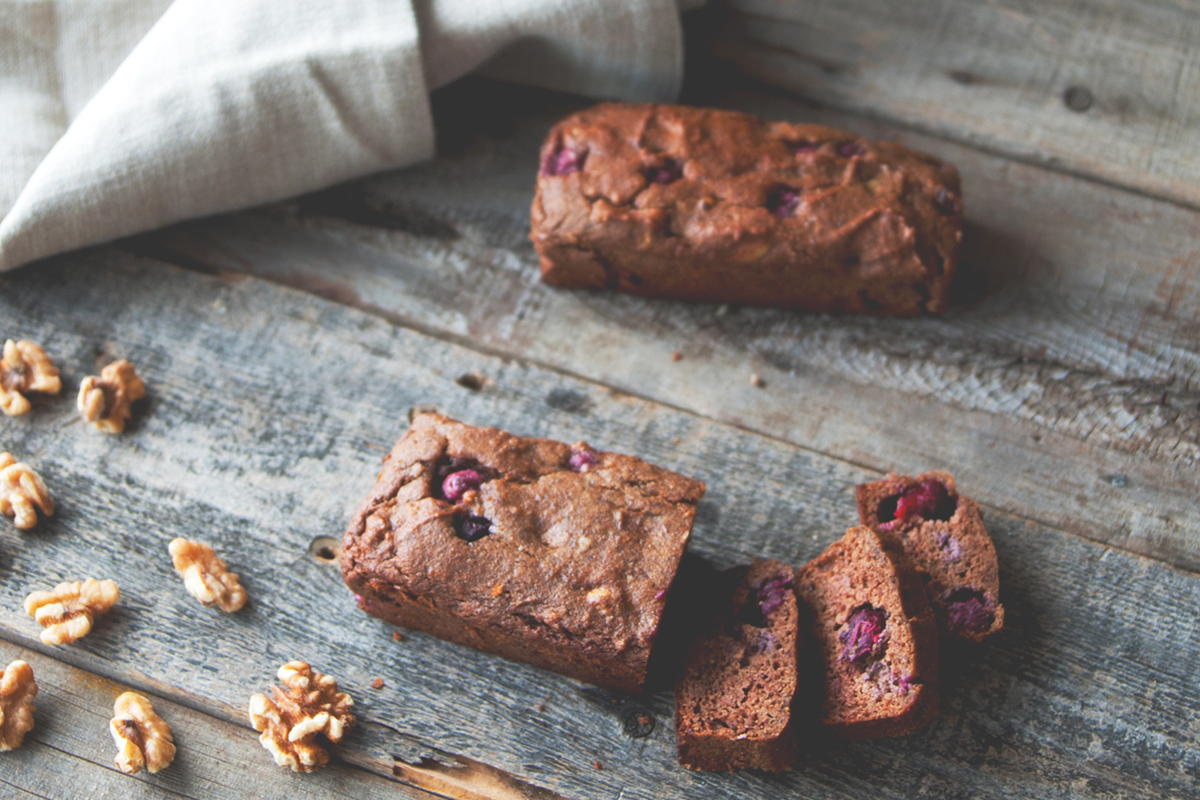 A loaf of berry and nut bread on a rustic wooden surface, with three slices cut and displayed in front. Scattered walnuts and a folded beige cloth are also visible.