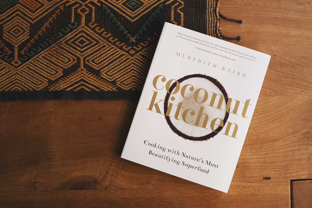 A cookbook titled Coconut Kitchen by Meredith Baird rests on a wooden table next to a woven textile. The book cover features a coconut ring and gold lettering.