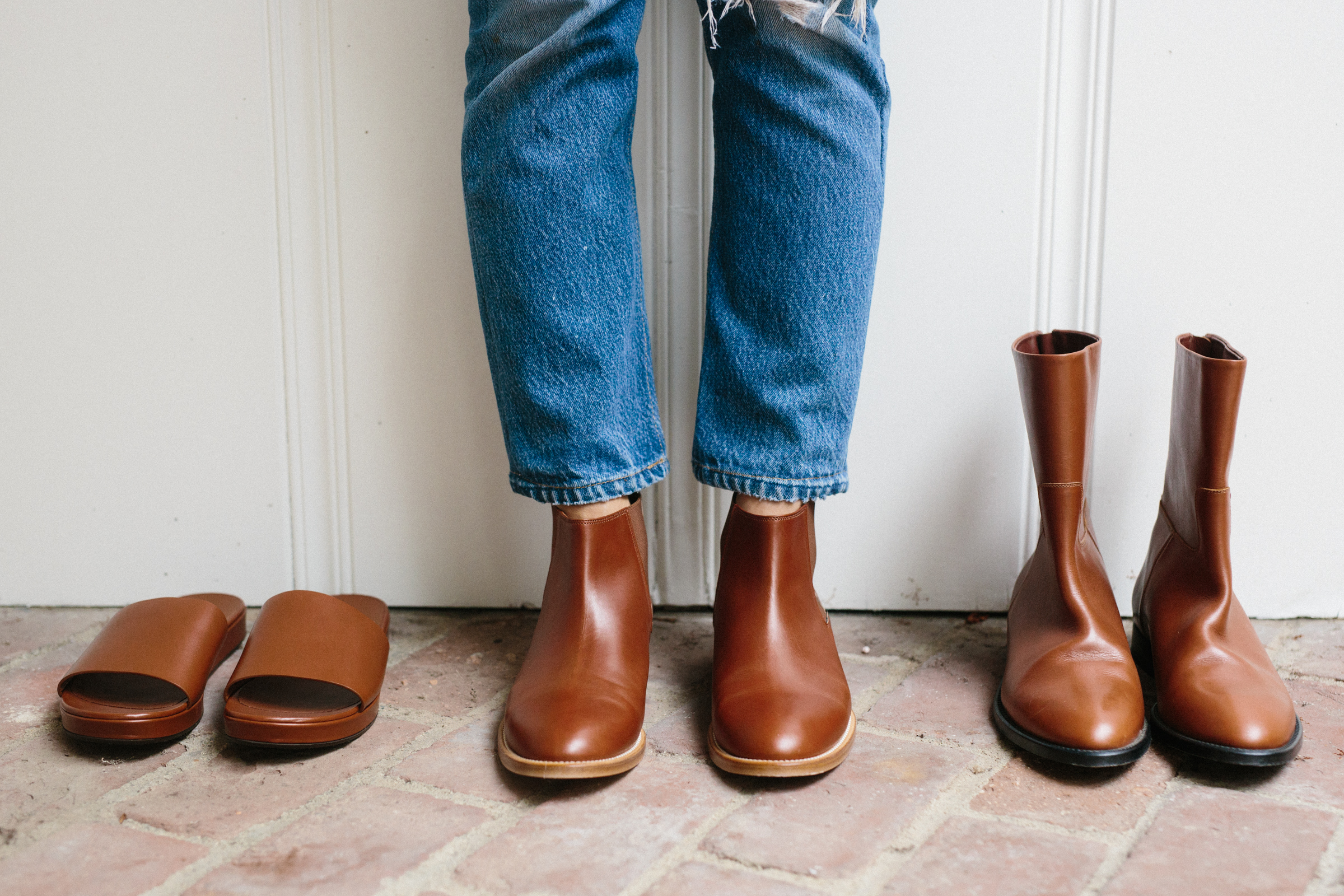 A person stands on a brick floor wearing blue jeans and brown leather saddle ankle boots. Next to them are a pair of brown leather slides on the left and taller brown leather boots on the right.