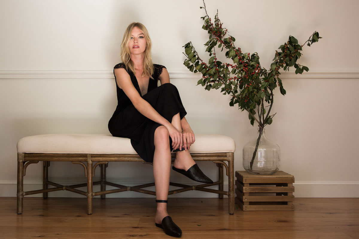 A woman in a black outfit sits on a cushioned bench against a white wall, next to a glass vase with leafy branches on a wooden crate. The minimalist setting with wooden flooring hints at quiet New Years Eve reflections.