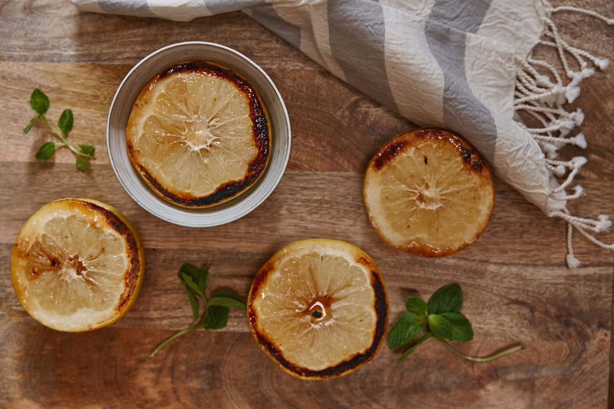 Four caramelized lemon halves are arranged on a wooden surface with a striped cloth, sprigs of mint, and a hint of grapefruit nearby; one lemon half is in a small metal dish.