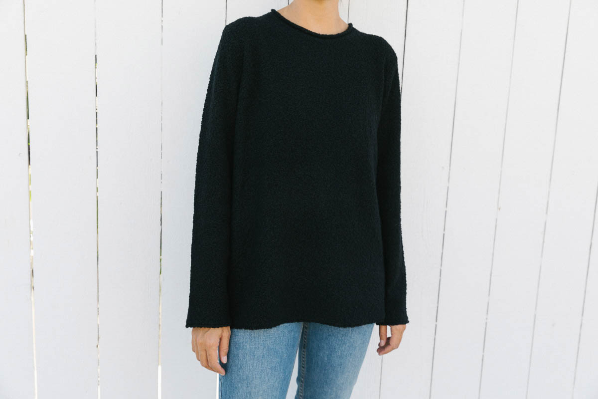 A person wearing a loose-fitting black long-sleeve sweater and blue jeans stands in front of a white wooden fence, with three fingers casually visible by their side. The persons head is partially out of frame.