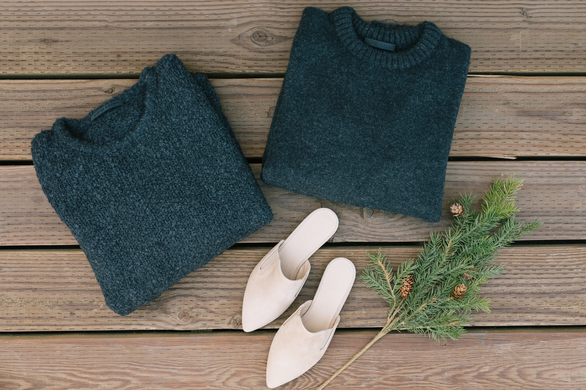 Two dark gray folded sweaters, a pair of beige slip-on shoes, and a small pine branch with pinecones are arranged on a wooden surface, capturing cozy vibes perfect for the holiday season.