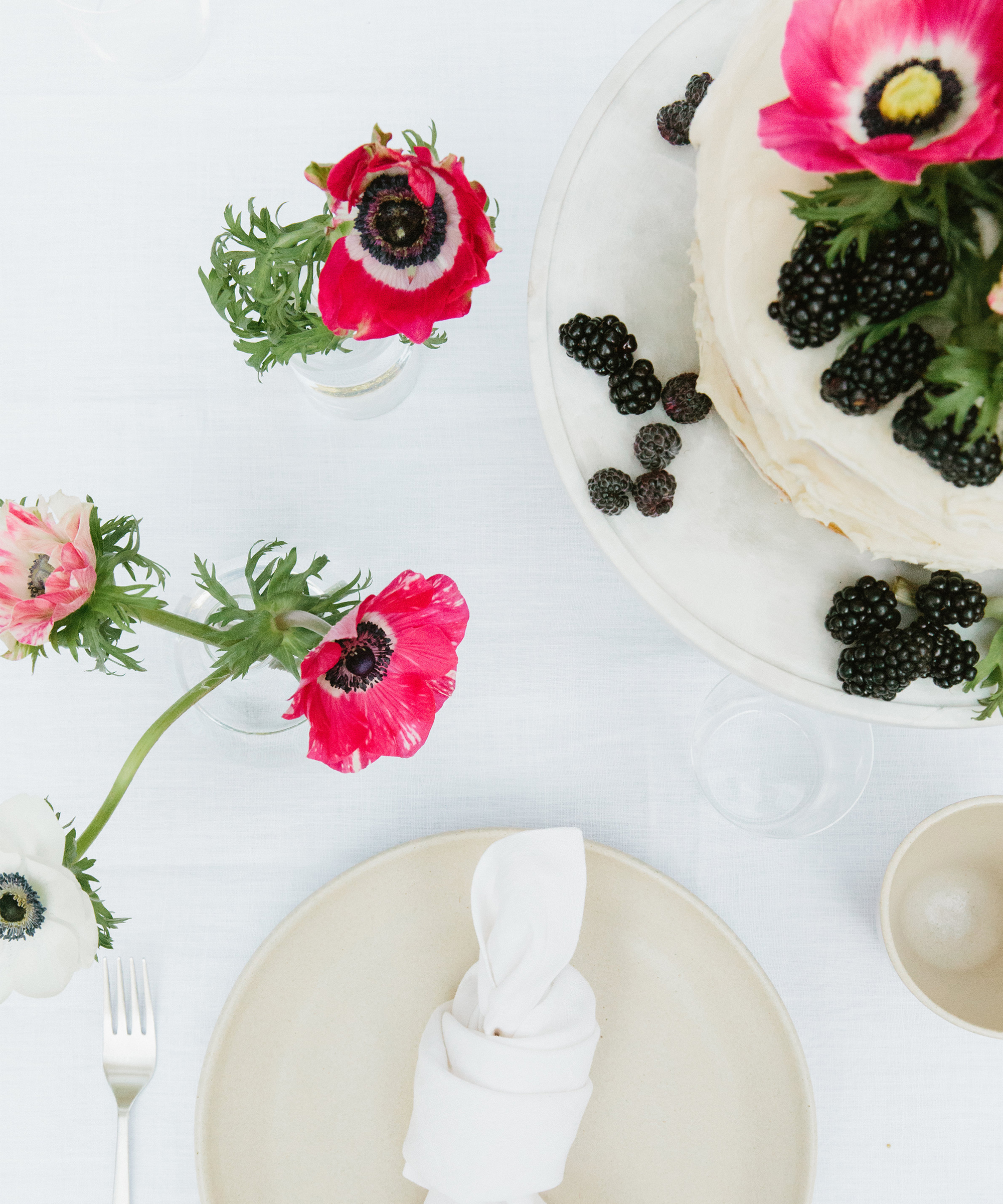 A tea-inspired table setting with a beige plate and napkin, a fork, small vases of pink and white anemone flowers, and a cake adorned with blackberries and bright flowers on a crisp white tablecloth.