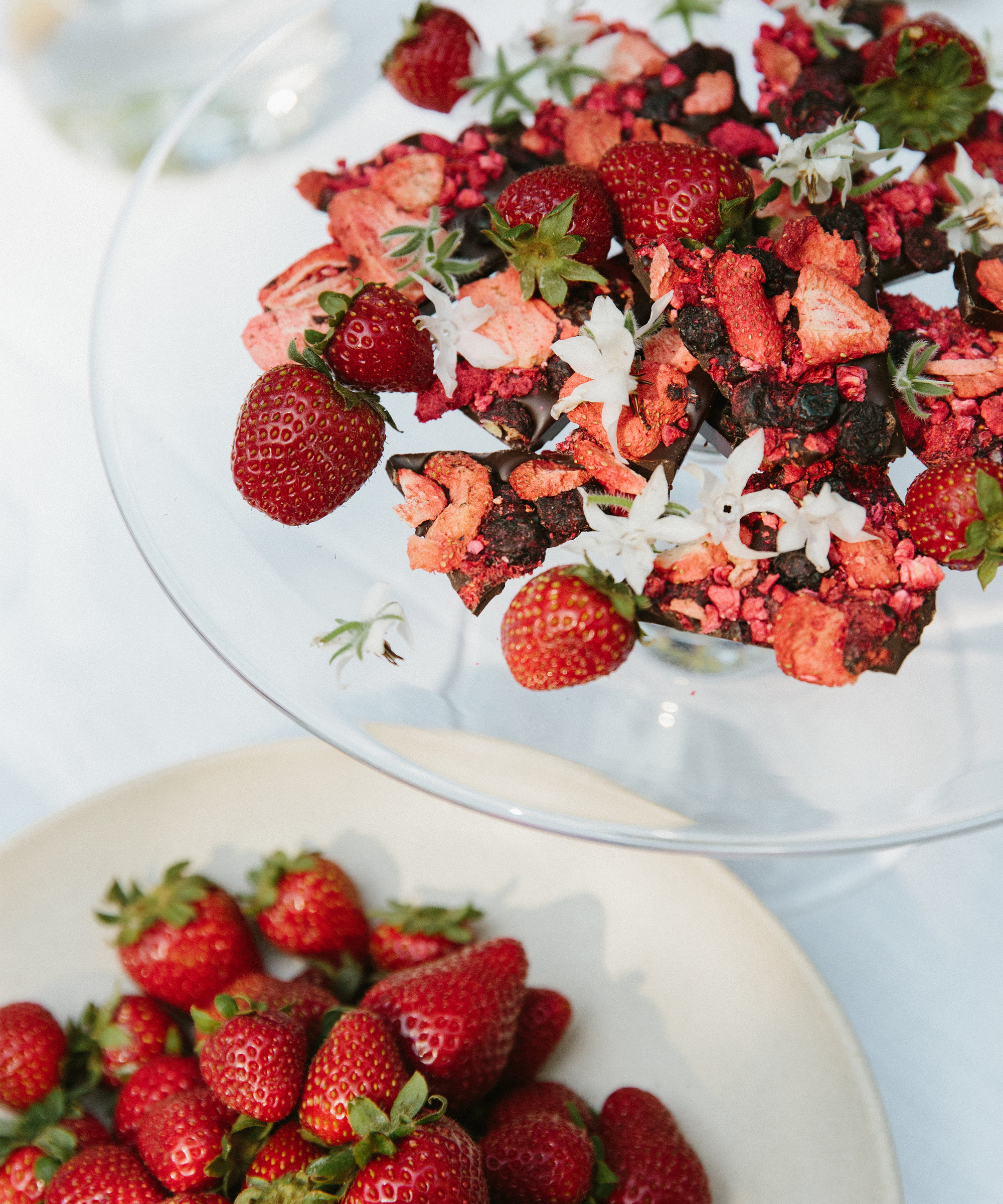 A glass dish holds whole strawberries, dried berries, chocolate pieces, and white edible flowers—perfect to pair with a cup of tea. Below, a beige bowl brims with fresh strawberries. Both dishes rest on a white surface.