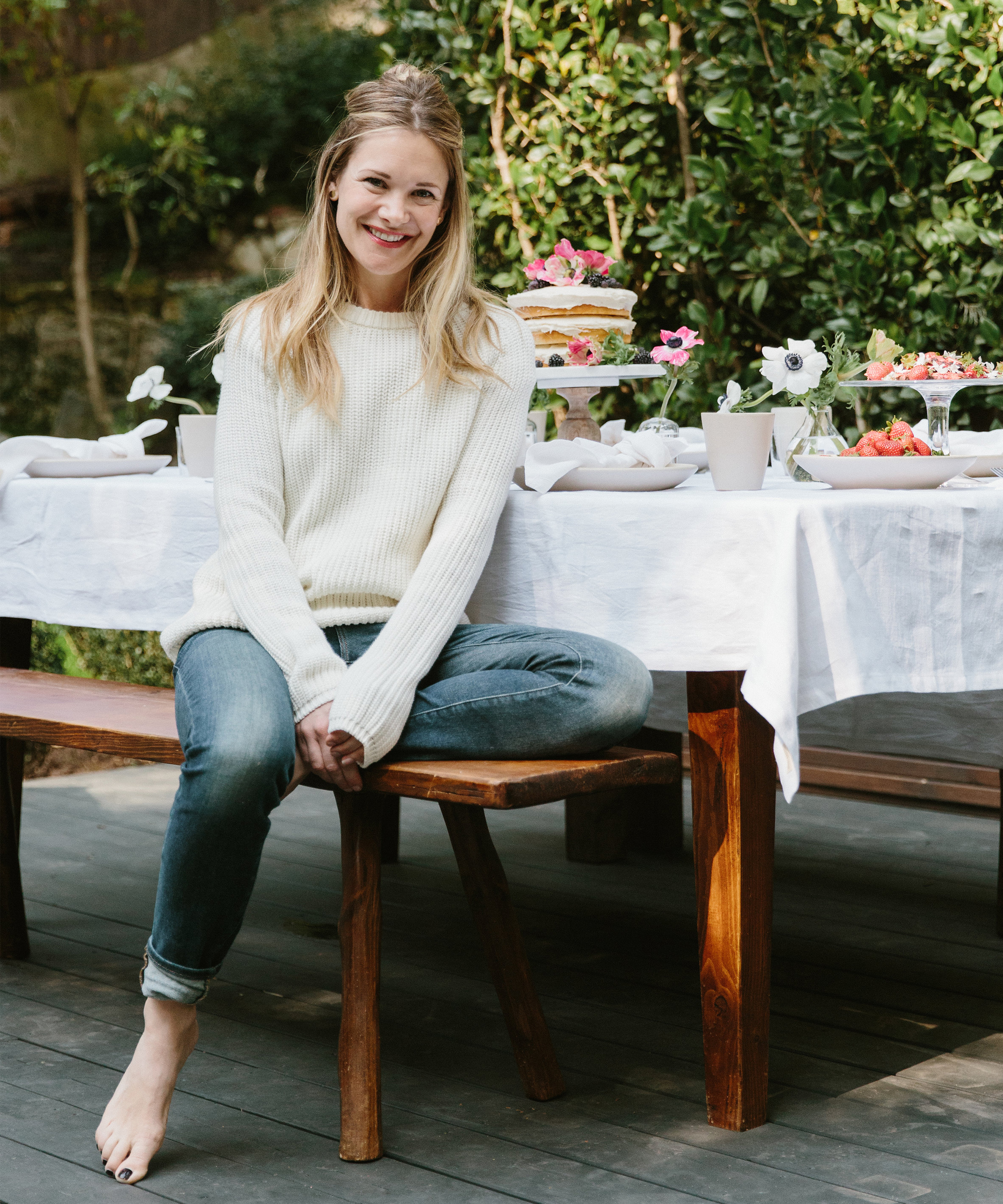 A woman in a white sweater and jeans sits barefoot on a wooden bench next to a table set with tea, flowers, cake, and dishes, outdoors on a patio with greenery in the background.