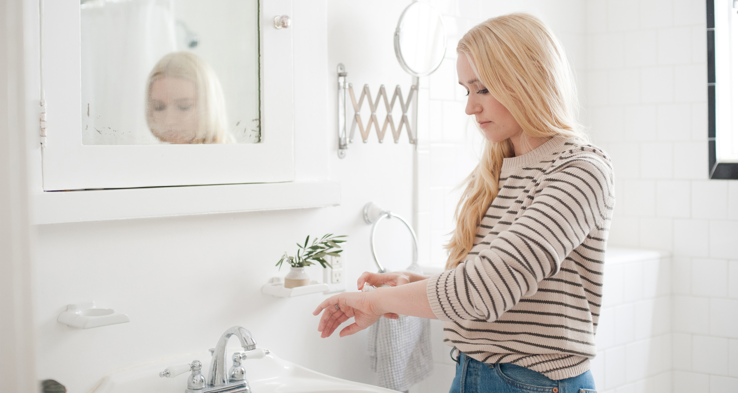 A woman with long blonde hair, wearing a striped sweater, stands in a bright bathroom applying lotion and oils to her hands at the sink. The white interior features a mirror and a small plant on a shelf.