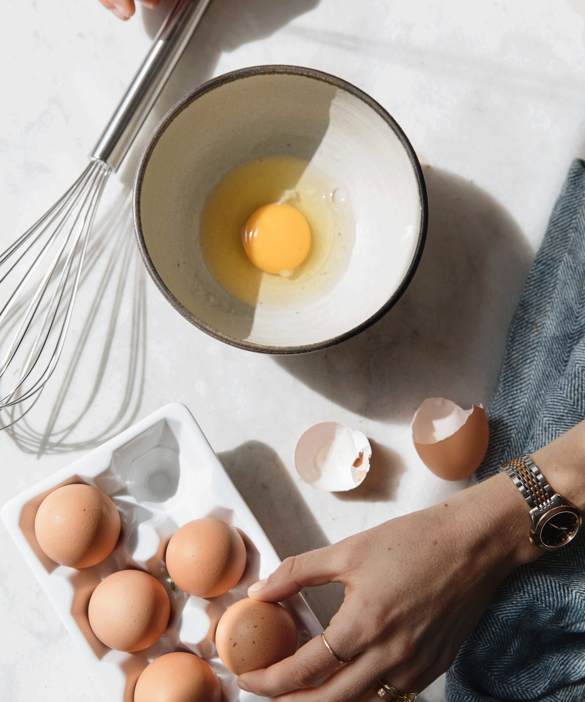 A hand holds an egg above a tray of eggs, with a cracked egg and shell in a bowl nearby. A metal whisk and a blue cloth are also on the white surface.