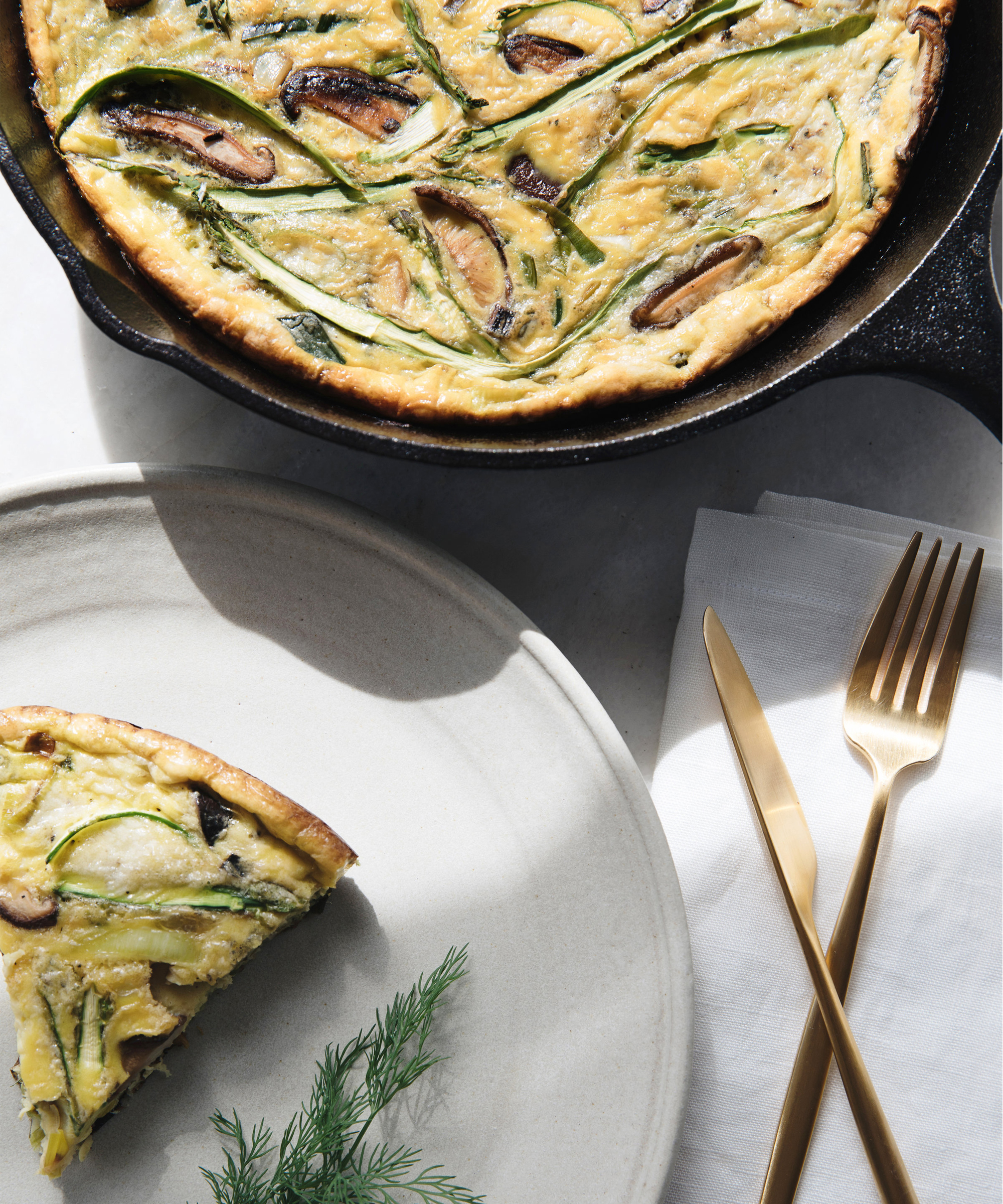 A slice of vegetable frittata with mushrooms and asparagus is served on a beige plate beside a gold fork and knife on a white napkin; the rest of the frittata sits in a cast iron skillet.