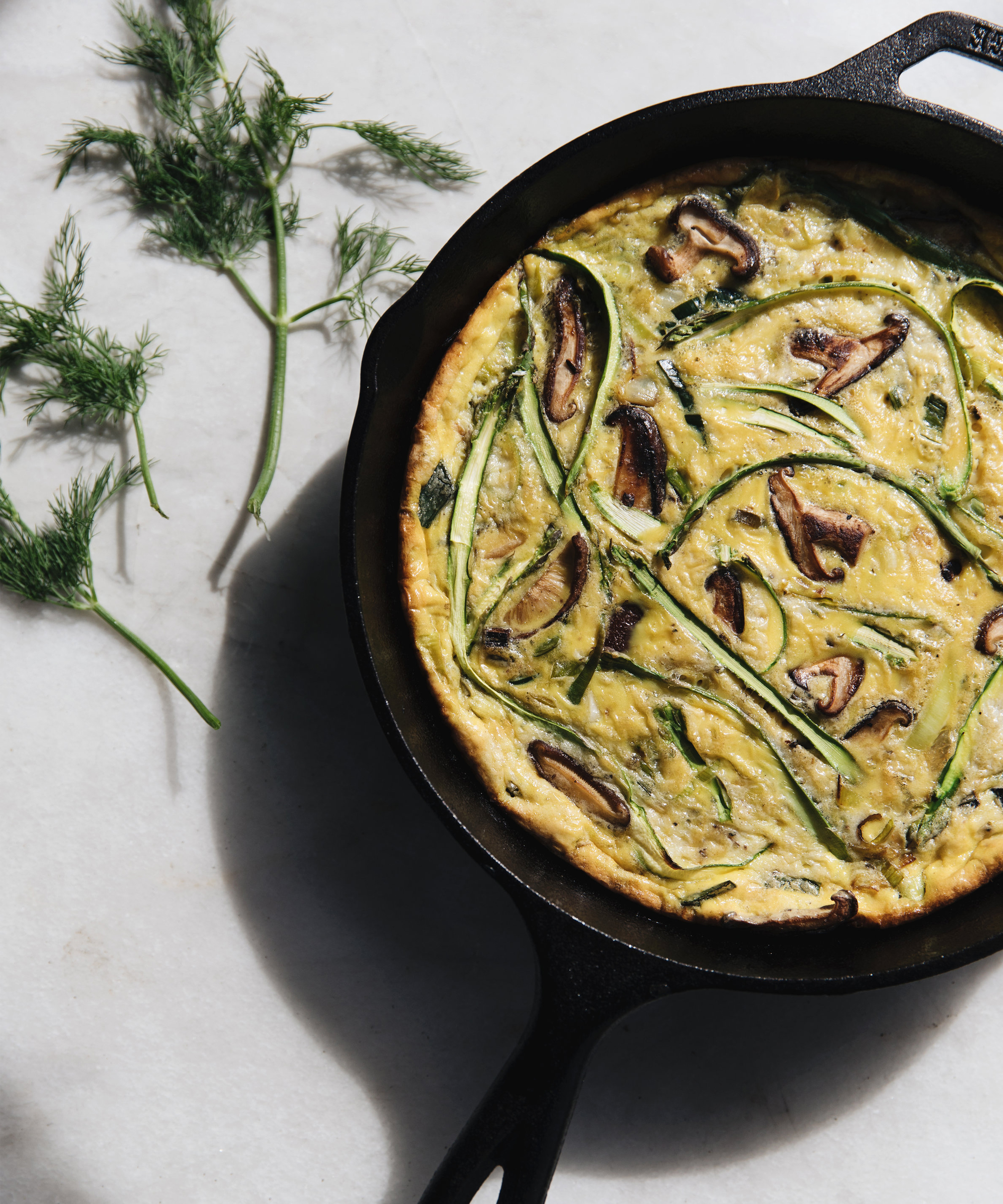 A frittata with mushrooms and green vegetables in a black cast iron skillet sits on a light-colored surface, with fresh dill sprigs placed beside it.