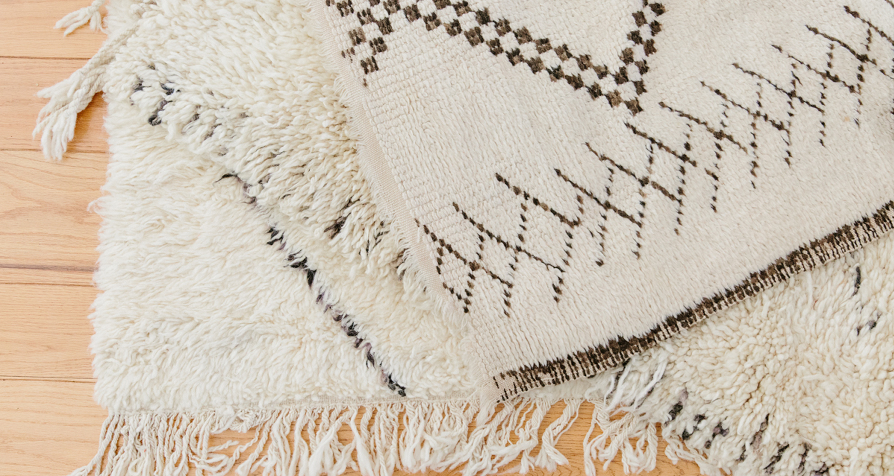 Two cream-colored rugs with fringe edges are layered on a light wooden floor. One rug is shaggy and fluffy, while the other showcases a flat weave with geometric black and brown patterns for added style.