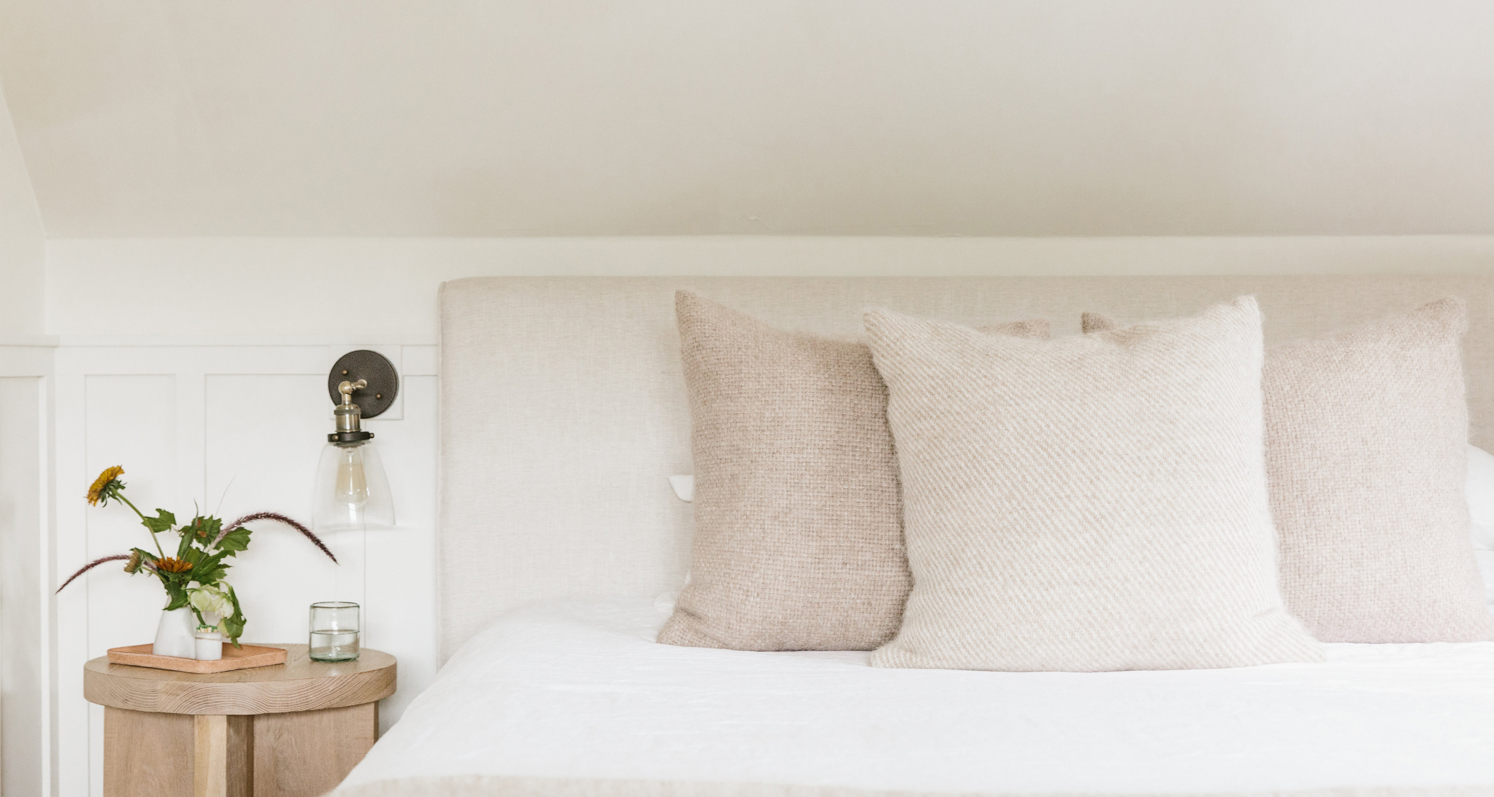 A neatly made bed with three large beige pillows, a white comforter, and a round wooden nightstand holding a small vase of flowers, a book, and a glass—complete with a designer wall-mounted lamp above the nightstand.