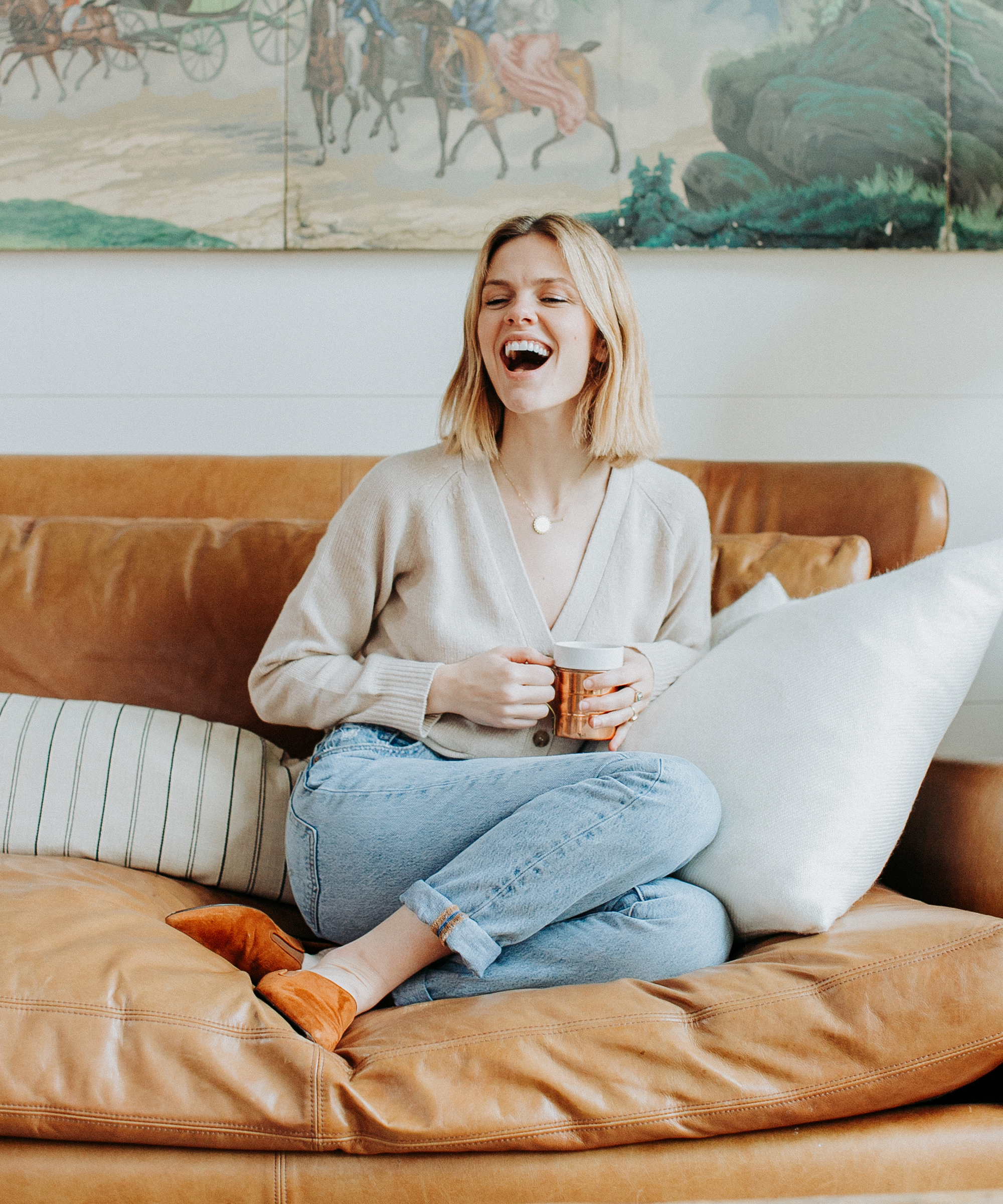 A woman resembling Brooklyn Decker with shoulder-length blonde hair sits on a tan leather couch, holding a mug and laughing. She wears a beige sweater, light blue jeans, and brown slippers. Behind her is a painting with horses and trees.